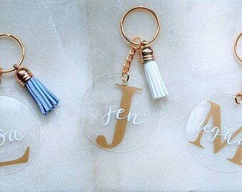 Op de afbeelding: Drie transparante acryl sleutelhangers met gouden letters en kwastjes. De sleutelhangers hebben de namen "Lisa", "Jen" en "Meghan" met hun bijbehorende beginletters in goud.