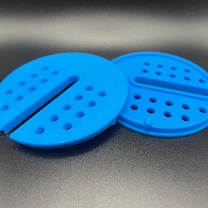 May include: Two bright blue, semi-circular objects with a series of small holes and a central slot. The objects are made of a solid material and appear to be 3D-printed. They are resting on a dark surface.