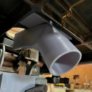 May include: A gray plastic dust collection attachment for a table saw. The attachment has a round opening and a rectangular base.
