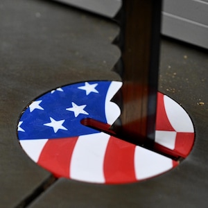 May include: A close-up of a circular saw blade cutting through a red, white, and blue American flag-themed safety cover. The cover is made of plastic and has a hole in the center for the blade to pass through.