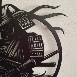 K&ouml;nnte beinhalten: Schwarze Metall-Wandkunst, die einen Samurai-Krieger im Profil zeigt. Das Kunstwerk zeigt detaillierte Darstellungen des Helms, der R&uuml;stung und eines Katana. Das Design umfasst einen kreisf&ouml;rmigen Rahmen und dekorative Elemente.