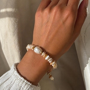 Könnte beinhalten: Ein handgefertigtes Armband mit einer großen, cremefarbenen Perle und kleinen, unregelmäßig geformten Muschelteilen. Das Armband ist an einer zarten Goldkette befestigt und hat einen kleinen Anhänger. Das Armband befindet sich an einem Handgelenk.
