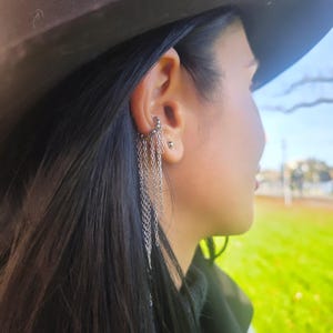 Könnte beinhalten: Silberner Ohrschmuck mit mehreren herabhängenden Ketten. Der Ohrschmuck wird am oberen Ohr getragen und fällt mit zarten Ketten herab. Die Person auf dem Bild trägt einen dunkelbraunen Hut und hat lange, dunkle Haare.