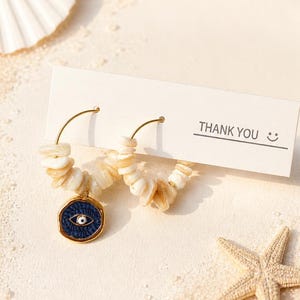 Könnte beinhalten: Goldene Creolen mit cremefarbenen Muschel-Akzenten und einem blauen und goldenen Evil-Eye-Anhänger. Die Ohrringe werden auf einer sandigen Oberfläche mit einem Seestern, Muscheln und einer Karte mit "DANKE" präsentiert.
