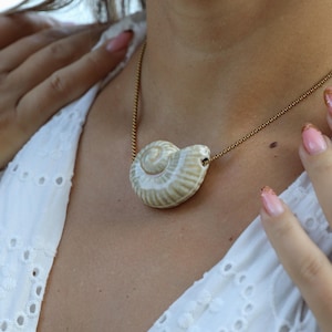 Könnte beinhalten: Eine Muschel-Anhänger-Halskette mit einer goldenen Kette. Der Anhänger ist eine cremefarbene und hellbraune Muschel mit detaillierten Rippen. Die Halskette wird über einem weißen Oberteil mit Lochmuster getragen.