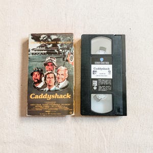 May include: A Caddyshack VHS tape and its cardboard sleeve. The sleeve features a graphic illustration of four men, a gopher, and a golf course with the text Caddyshack. The black plastic tape has a Warner Home Video label with the title Caddyshack.