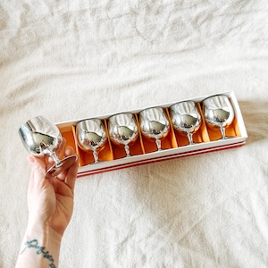 May include: A set of six silver-colored brandy snifters in a red and white box with an orange interior. One glass is held by a hand. The glasses have a rounded bowl and a short stem. The box is on a white surface.