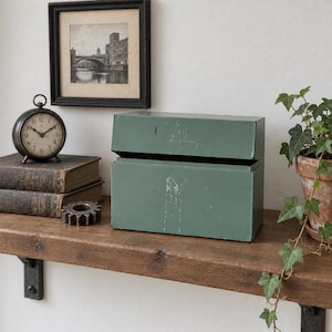 May include: A rectangular, sage green metal storage box with a hinged lid. The front of the box features a faint circular stamp and the text "MADE IN CANADA" embossed on the upper portion, with visible surface wear and paint scuffs.