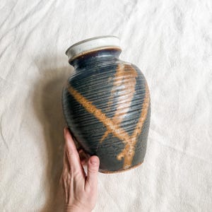 May include: A ceramic vase with a dark charcoal gray body featuring horizontal ribbed textures and a prominent diagonal tan glaze streak. The vase has a smooth, light-colored rim and a rounded, bulbous shape that tapers toward the top opening.