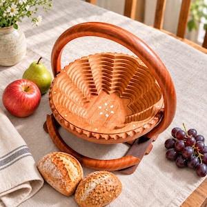 May include: A collapsible wooden fruit basket with a circular handle and a carved flower design at the base. The basket features a lattice-like structure that expands from a flat disc into a bowl shape, crafted from polished reddish-brown wood.