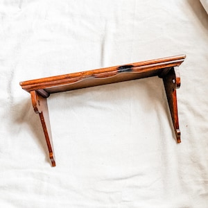 May include: A wall-mounted wooden shelf featuring a medium brown finish with a visible wood grain. The shelf has a flat rectangular top surface and two downward-pointing side brackets with curved edges and circular cutouts for mounting.