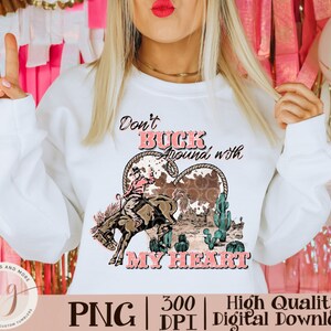 May include: White sweatshirt with a graphic design featuring a cowboy riding a bucking bronco inside a heart shape. The text reads "Don't Buck Around with My Heart". The design includes cacti and a cowhide pattern.