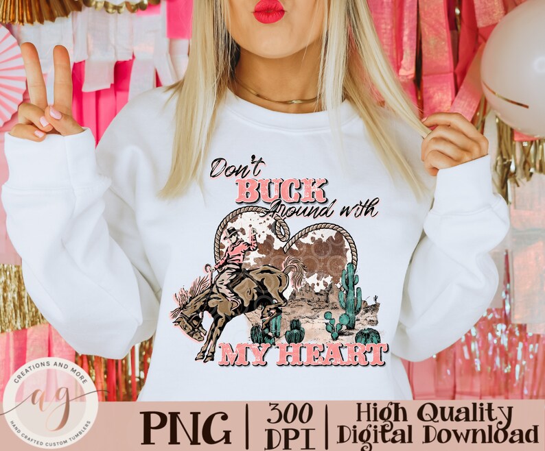 May include: White sweatshirt with a graphic design featuring a cowboy riding a bucking bronco inside a heart shape. The text reads "Don't Buck Around with My Heart". The design includes cacti and a cowhide pattern.
