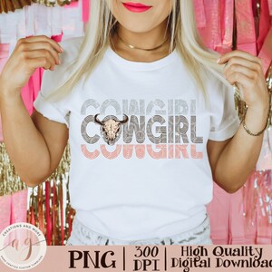 May include: A white t-shirt with the text "COWGIRL" repeated three times in a retro style font. The word "COWGIRL" is layered with a skull and crossbones graphic in the center. The text is in shades of gray and pink.