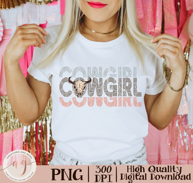 May include: A white t-shirt with the text "COWGIRL" repeated three times in a retro style font. The word "COWGIRL" is layered with a skull and crossbones graphic in the center. The text is in shades of gray and pink.