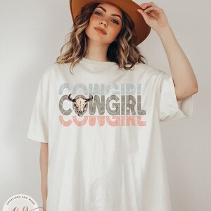 May include: A white t-shirt with a graphic design of the word "COWGIRL" in a retro style with a cow skull in the center. The design is in shades of blue, pink, and brown.