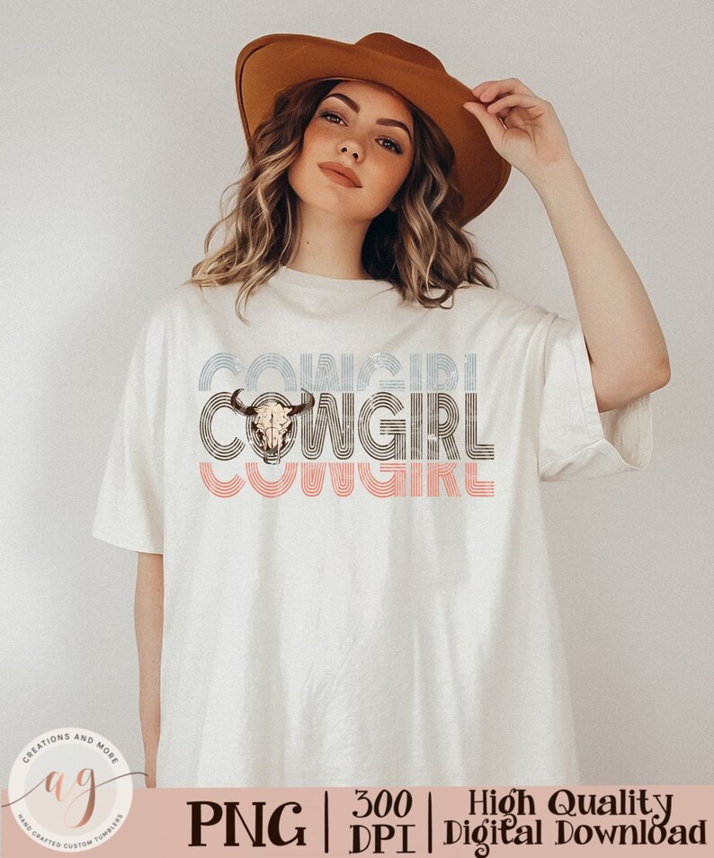 May include: A white t-shirt with a graphic design of the word "COWGIRL" in a retro style with a cow skull in the center. The design is in shades of blue, pink, and brown.