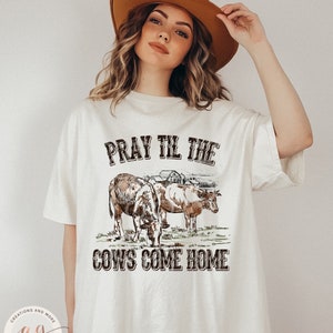 May include: A white t-shirt with a brown graphic design of two cows in a field with a farmhouse in the background. The text "Pray Til The Cows Come Home" is printed in brown on the shirt.