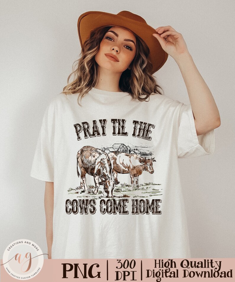 May include: A white t-shirt with a brown graphic design of two cows in a field with a farmhouse in the background. The text "Pray Til The Cows Come Home" is printed in brown on the shirt.