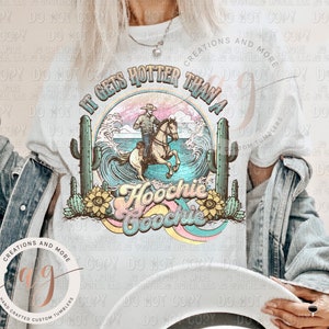 May include: A white t-shirt with a graphic design featuring a cowboy riding a horse through a wave with the text "It gets hotter than a Hoochie Coochie". The design also includes cacti and sunflowers.