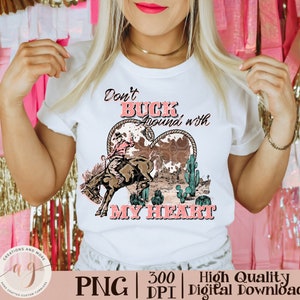May include: White t-shirt with a graphic featuring a rodeo cowboy riding a bucking bronco inside a heart-shaped design. The text "Don't Buck Around with My Heart" is displayed in pink and black.