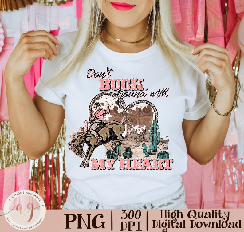 May include: White t-shirt with a graphic featuring a rodeo cowboy riding a bucking bronco inside a heart-shaped design. The text "Don't Buck Around with My Heart" is displayed in pink and black.