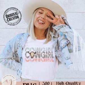 May include: A white t-shirt with a graphic design of the word "COWGIRL" repeated three times in a retro style with a cow skull in the center. The shirt is being worn by a person with blonde hair and a white cowboy hat.