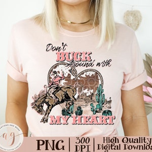 May include: A light pink t-shirt featuring a graphic design with the text "Don't Buck Around with My Heart." The design includes a cowboy riding a bucking bronco, cacti, and a heart-shaped element.