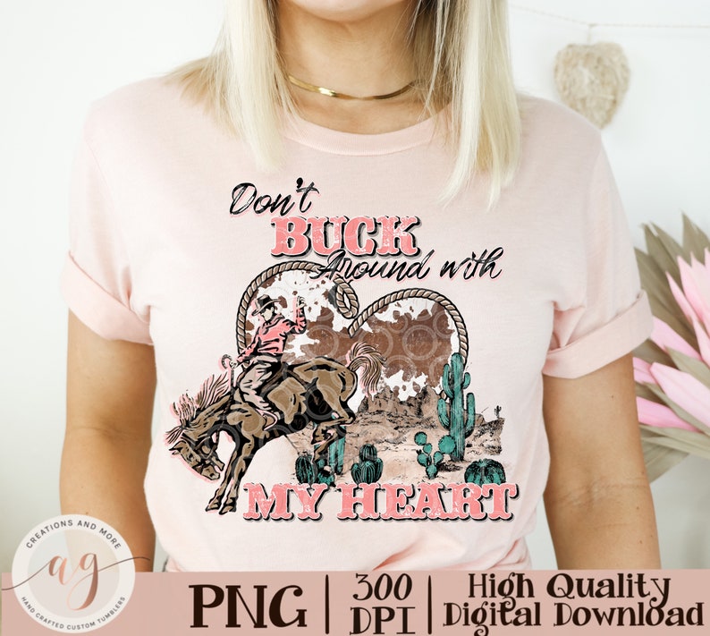 May include: A light pink t-shirt featuring a graphic design with the text "Don't Buck Around with My Heart." The design includes a cowboy riding a bucking bronco, cacti, and a heart-shaped element.