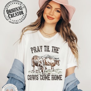 May include: A white t-shirt with the text "Pray Til The Cows Come Home" in black lettering. The design features two brown cows in a field with a farmhouse in the background. The shirt is worn by a person wearing a pink cowboy hat.