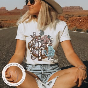 May include: White t-shirt featuring a floral design and the text "He is Risen" within a brown rope circle. The design also incorporates two crosses and butterflies.