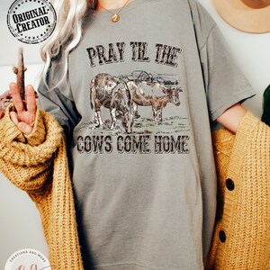May include: A gray t-shirt with the text "Pray Till The Cows Come Home" in black. The design features a vintage illustration of two cows in a field.