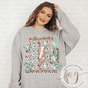 May include: A gray sweatshirt with a graphic design of the word "Baseball Mama" in a distressed font with a lightning bolt and cactus accents. The design is in shades of teal, brown, and white.