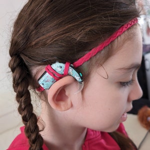 Può includere: Una giovane ragazza con i capelli intrecciati indossa una fascia rosa e un apparecchio acustico con un design colorato. L'apparecchio acustico è attaccato a una fascia con una piccola luce verde.