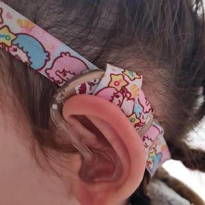 Könnte beinhalten: Ein beiges Hinter-dem-Ohr-Hörgerät, das mit einem hellrosa Stoffhalteband gesichert ist, welches ein wiederkehrendes Muster aus Zeichentrickfiguren, Sternen und Einhörnern zeigt. Das Gerät verfügt über einen transparenten Kunststoffschlauch.