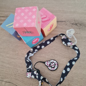 Puede incluir: Un conjunto de bloques de madera coloridos con diferentes patrones de animales y geométricos. Los bloques están apilados uno encima del otro. Una correa con estampado de margaritas blancas y negras con un pequeño dispositivo blanco y negro también está en la imagen.