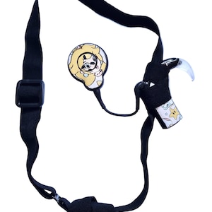 Può includere: Un cinturino elastico nero con fibbia regolabile, dotato di un ciondolo circolare con l'illustrazione di un personaggio dei cartoni animati e un attacco secondario decorato con un motivo di stelle gialle e piccole forme bianche su sfondo chiaro.