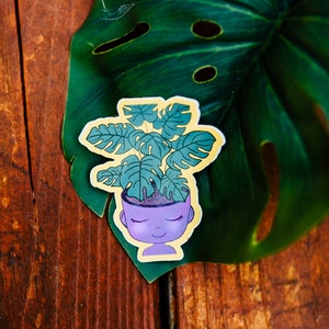 Pode incluir: Um autocolante com uma ilustração de desenho animado de uma pessoa com uma planta crescendo em sua cabeça. A planta tem folhas grandes e verdes. A cabeça da pessoa é roxa e o autocolante tem uma borda amarela. O autocolante está em uma folha verde.