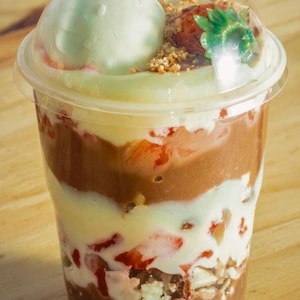 May include: A clear plastic cup filled with layers of chocolate pudding, white cream, strawberries, and a scoop of green ice cream topped with a strawberry and a sprinkle of granola.