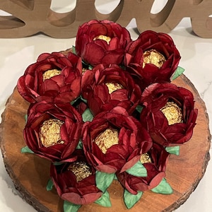 Op de afbeelding: Een ronde houten schijf met een cluster van bordeauxrode papieren bloemen met daarin chocoladebonbons verpakt in goudfolie. De bloemen zijn in een cirkelvormige opstelling op de houten schijf geplaatst.