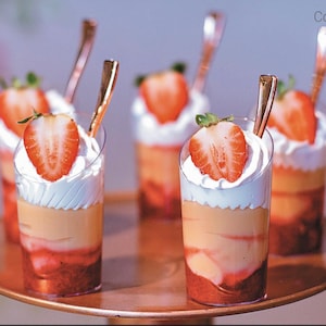 Puede incluir: Cuatro vasos de postre de vidrio transparente llenos de capas de crema batida, fruta y una crema amarilla. Los vasos están cubiertos con fresas frescas y están dispuestos en una bandeja de color cobre. El texto "Copo Verrine" es visible en la esquina superior derecha de la imagen.