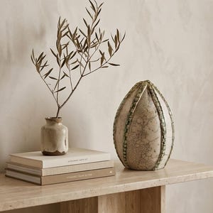 Puede incluir: Naturaleza muerta decorativa con un pequeño jarrón de cerámica beige con hojas secas, una pila de libros y un jarrón grande, texturizado, en forma de huevo con detalles verdes. Los objetos están colocados sobre una mesa de piedra clara.
