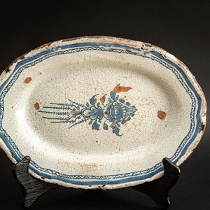 Può includere: Piatto ovale antico in ceramica con smalto craquelé bianco. Il piatto presenta un motivo floreale blu e un bordo blu. Ci sono anche piccole macchie marrone-arancio sulla superficie. Il piatto è esposto su un supporto nero.