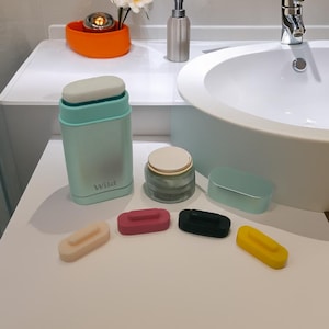 Peut inclure: Un ensemble de cinq couvercles de déodorant en silicone de forme ovale et de couleurs vives. Les couvercles sont dans des tons de rose, noir, jaune et beige. Les couvercles sont disposés en rangée sur une surface blanche. Un déodorant en plastique bleu clair avec le mot "Wild" imprimé dessus est en arrière-plan.