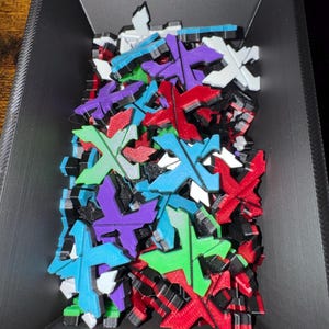May include: A collection of colorful, X-shaped objects in a black container. The objects are in various colors, including red, blue, green, purple, and white. The container is rectangular and has a textured surface.