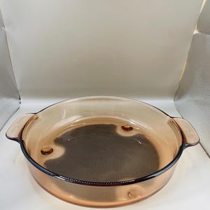 May include: A round, amber-coloured glass baking dish with two handles. The dish has a dark, textured interior and is designed for oven use. The glass is transparent, allowing visibility of the contents.