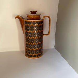 Puede incluir: Una cafetera alta de cerámica marrón con tapa y asa. El cuerpo presenta un patrón repetido de formas de diamantes negros y naranjas. El pico y el asa son de color marrón.