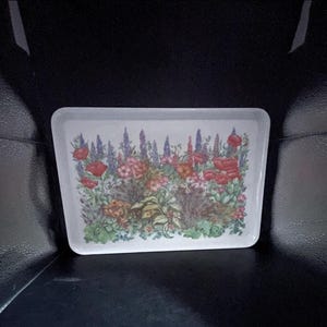 Pode incluir: Uma bandeja de servir retangular branca com uma ilustração botânica colorida de várias flores de jardim, incluindo papoulas vermelhas, espigas roxas e folhagem verde dispostas num padrão floral denso e em camadas no centro.