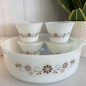 May include: A set of vintage white glass Pyrex bowls and containers. The large bowl and smaller containers feature a brown floral design. The set includes a large bowl with handles, two small bowls, and two lids.
