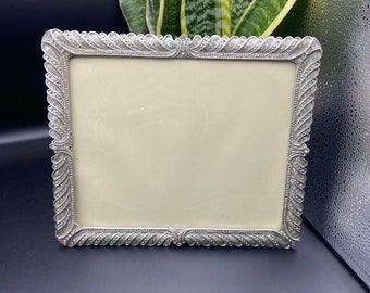 May include: A silver picture frame with a decorative, ornate border. The frame has a light beige mat and is empty.